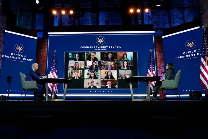 Biden and Harris, both wearing masks, sit on a stage behind desks and socially distanced, with a screen showing members of the task force in the background